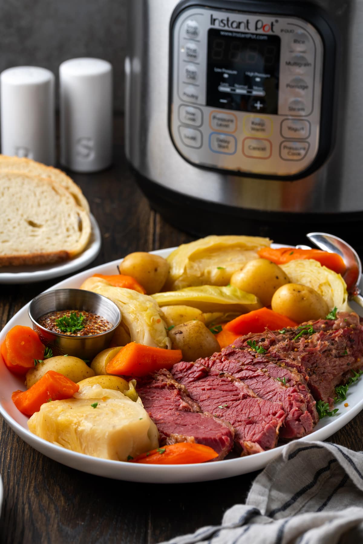 In Scheiben geschnittenes Corned Beef auf einem Teller neben Kohl, Kartoffeln und Karotten, mit dem Instant Pot und einer Reihe von Brotscheiben im Hintergrund.