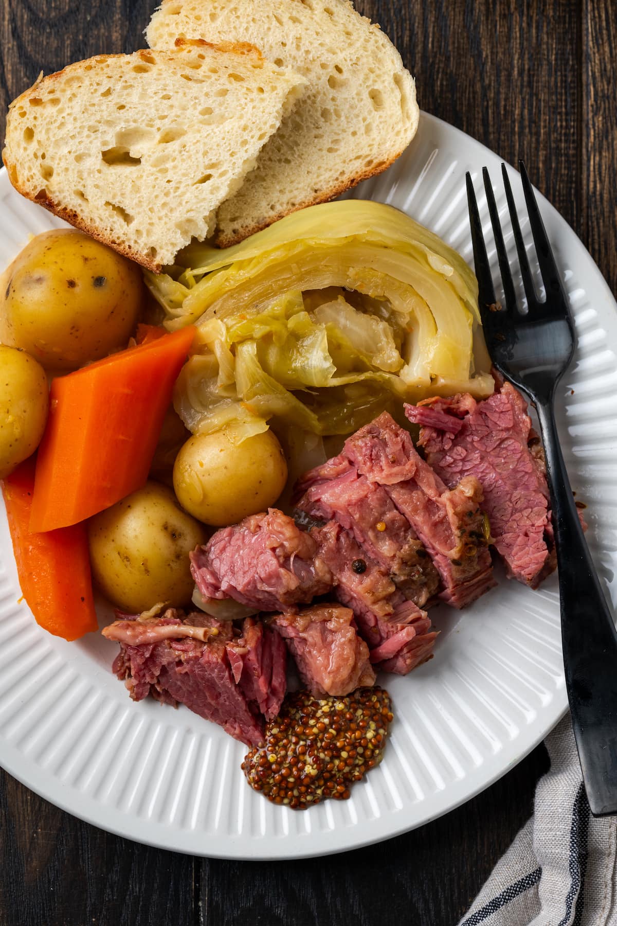 Draufsicht auf geschnittenes Corned Beef auf einem Teller mit Kohl, Kartoffeln, Karotten und Brotscheiben, daneben eine Gabel.