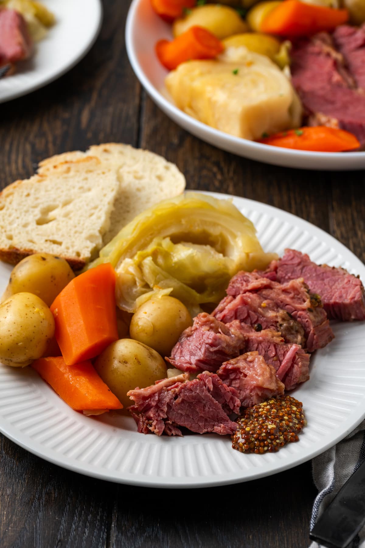 In Scheiben geschnittenes Corned Beef auf einem Teller neben Kohl, Kartoffeln, Karotten und Brotscheiben, mit einem zweiten Teller im Hintergrund.