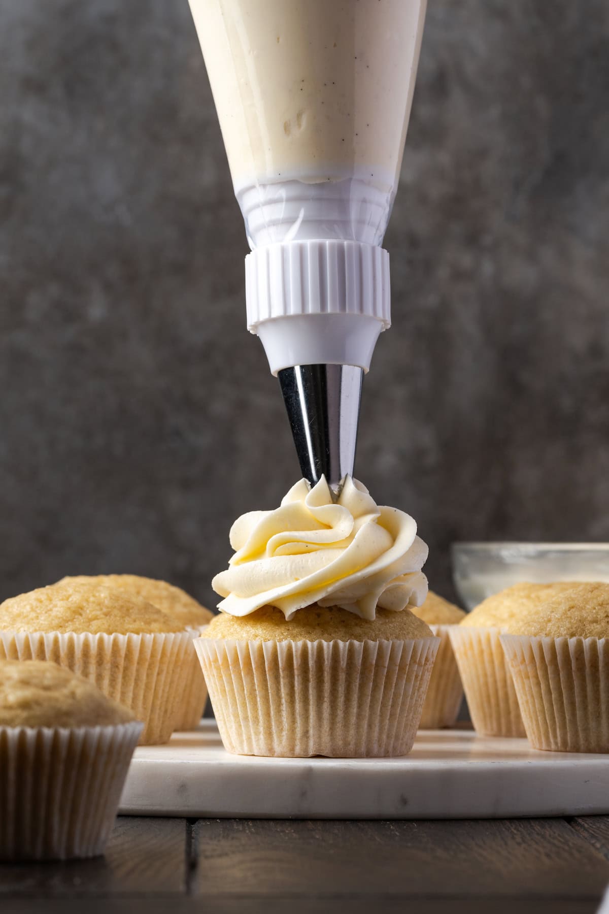 Ein Spritzbeutel mit einer Spritztülle, der einen Strudel aus Buttercreme auf einen Cupcake spritzt, mit weiteren ungefrosteten Cupcakes im Hintergrund.