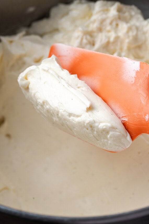 Ein orangefarbener Gummispatel, der einen Löffel Schweizer Baiser-Buttercreme über einer Schüssel mit Zuckerguss hält.