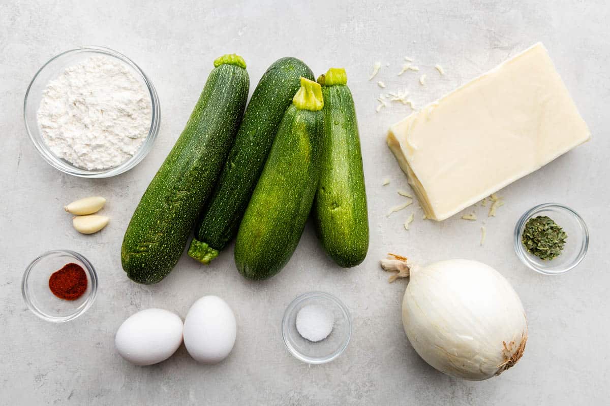 Ein Overhead-Bild von ganzen Zucchini, frischem Gemüse und einem Stück Käse mit Eiern sowie trockenen und feuchten Zutaten.