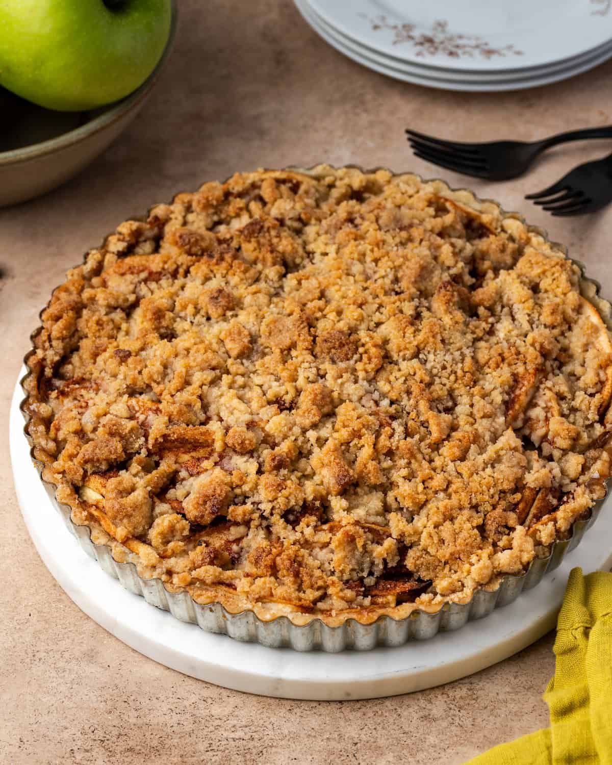 Gebackener Apfelstreuselkuchen in einer Tarteform mit einem Stapel Teller und einer Schüssel mit Äpfeln im Hintergrund.